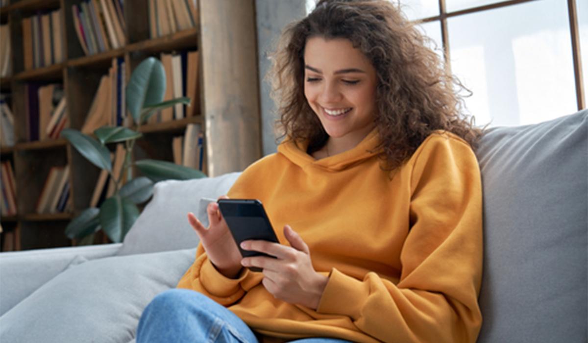 Donna sorridente seduta sul divano mentre consulta lo smartphone per gestire i dispositivi connessi della casa