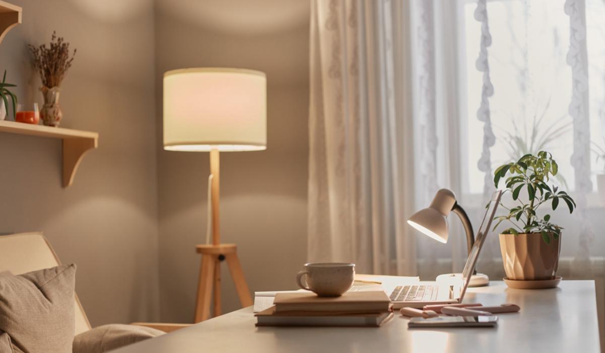 Salotto accogliente con lampada da terra accesa e scrivania illuminata da una lampada da tavolo, con laptop, tazza e pianta verde