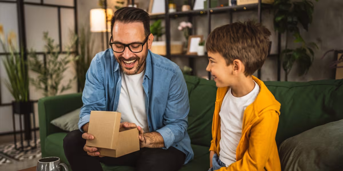 Uomo sorridente seduto sul divano apre un regalo mentre un bambino lo guarda con entusiasmo