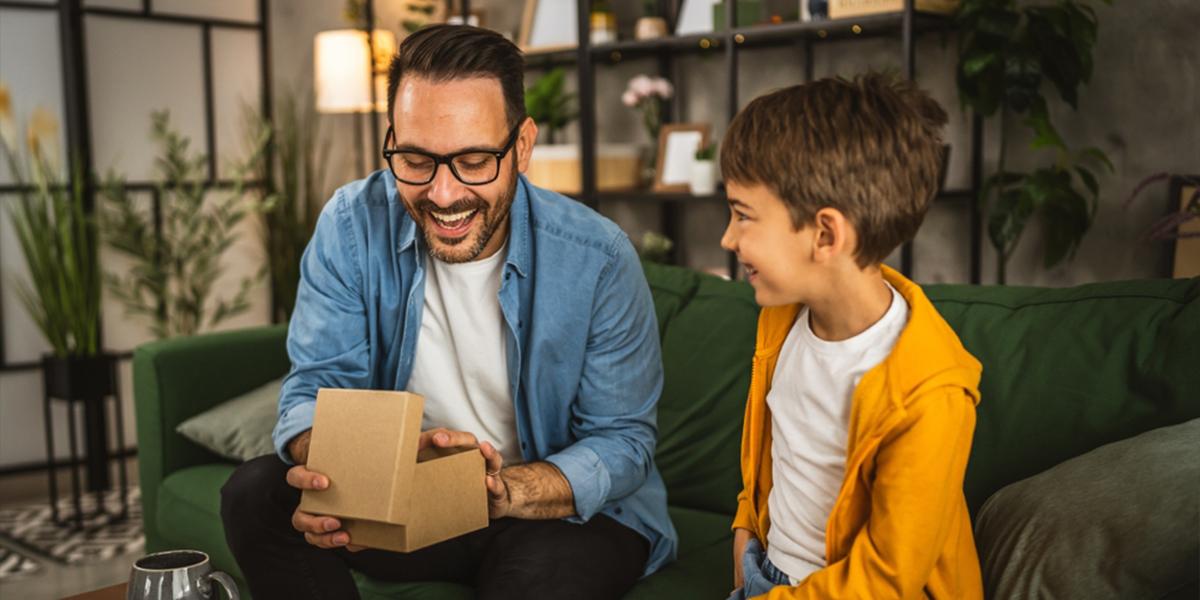Uomo sorridente seduto sul divano apre un regalo mentre un bambino lo guarda con entusiasmo
