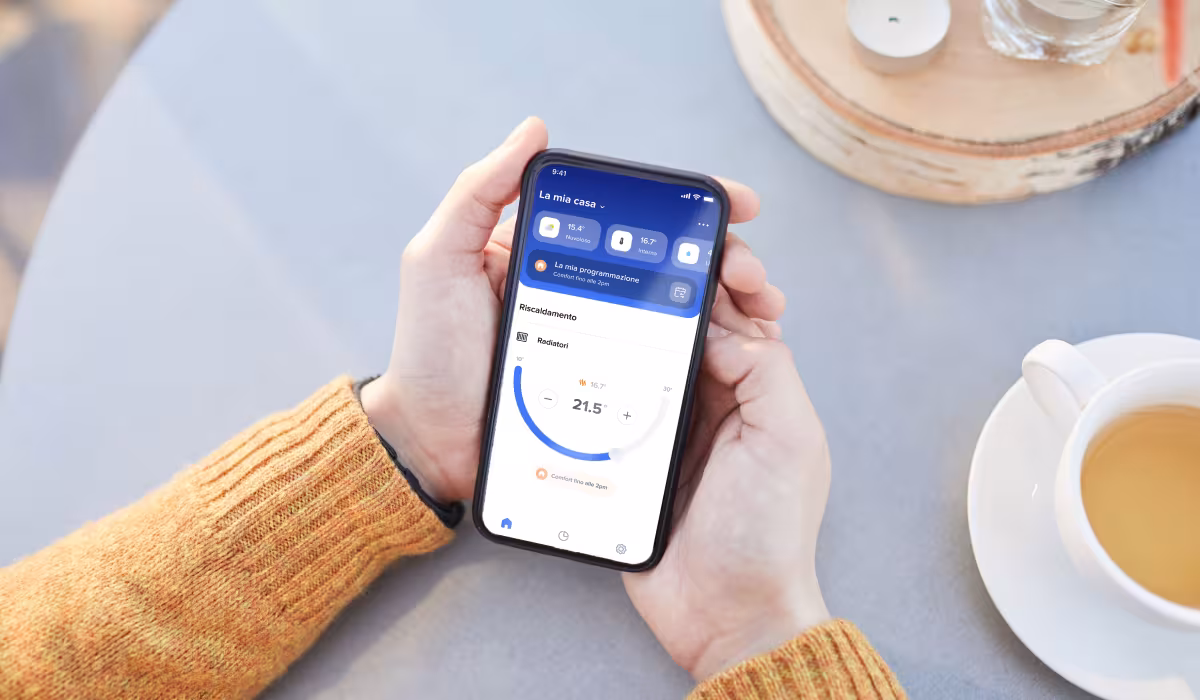Mani su un tavolo accanto a una tazza che tengono uno smartphone con aperta l’app Home + Control di BTicino per la gestione del riscaldamento
