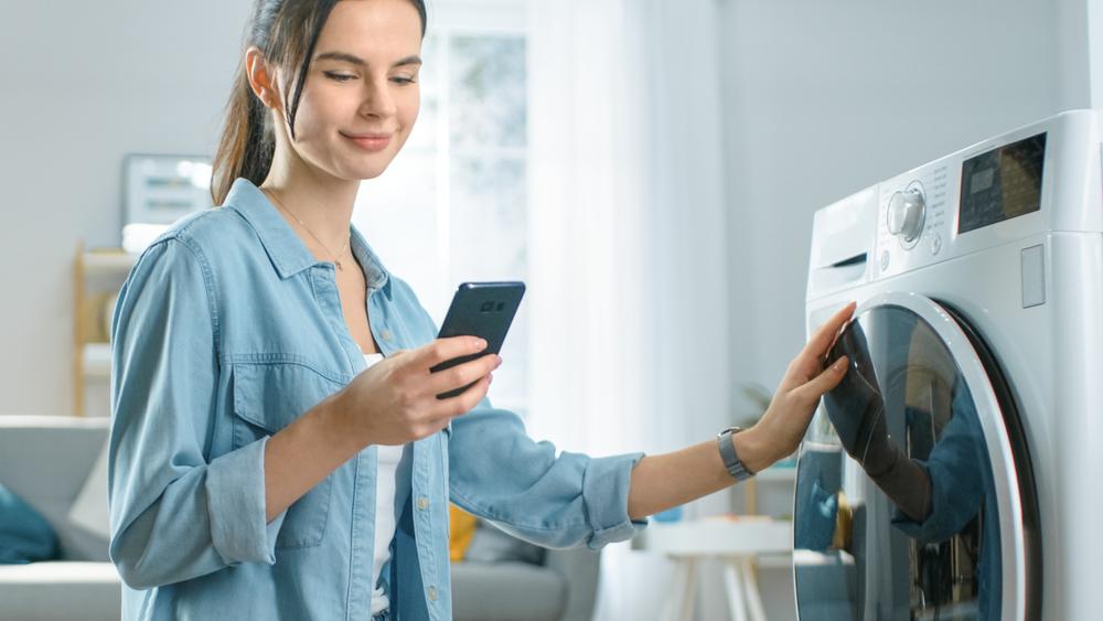 Donna accanto alla lavatrice mentre imposta il ciclo di lavaggio tramite il suo smartphone