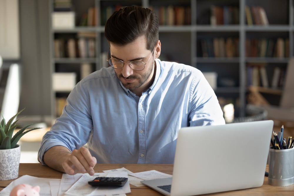 Uomo con gli occhiali seduto al tavolo mentre utilizza una calcolatrice per calcolare le spese mensili