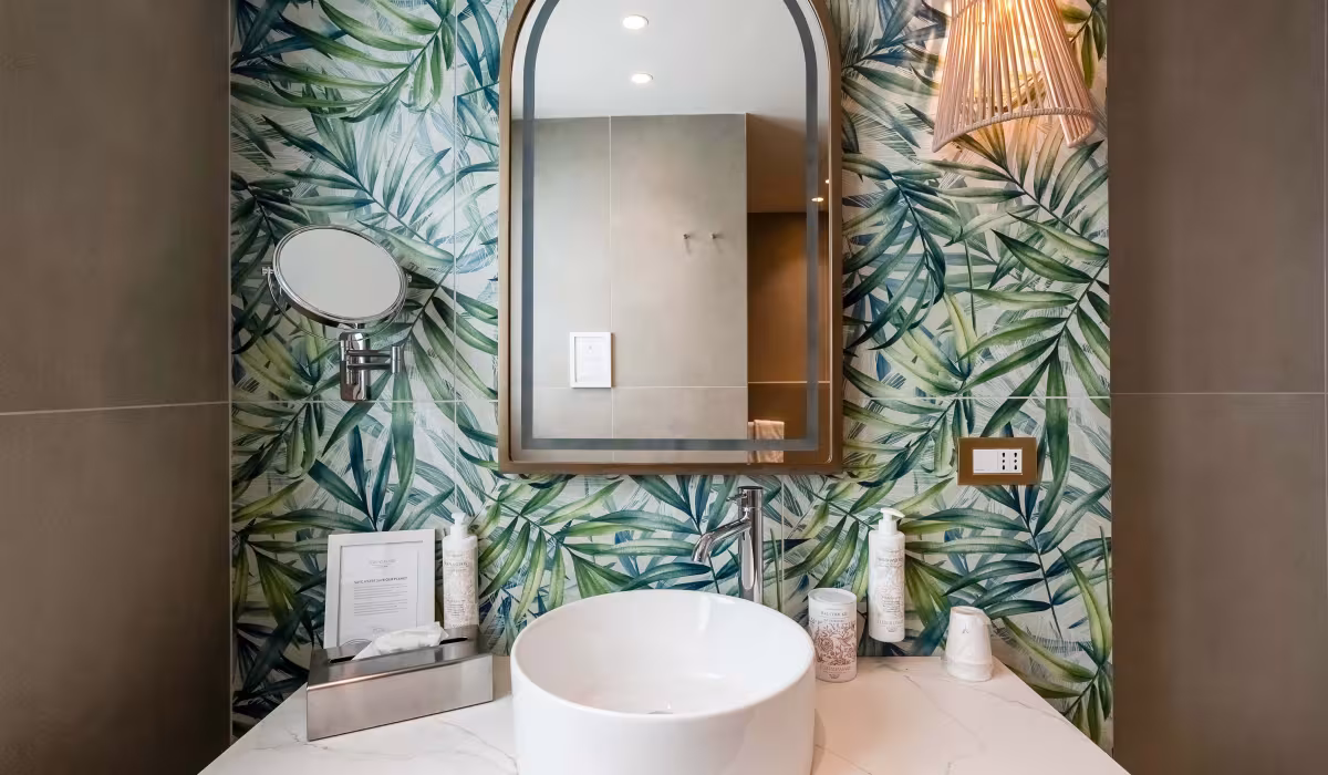 Bagno del Terradamari Resort & Spa con lavabo moderno, specchio ad arco, parete decorata con motivi tropicali e placche Livinglight Bticino