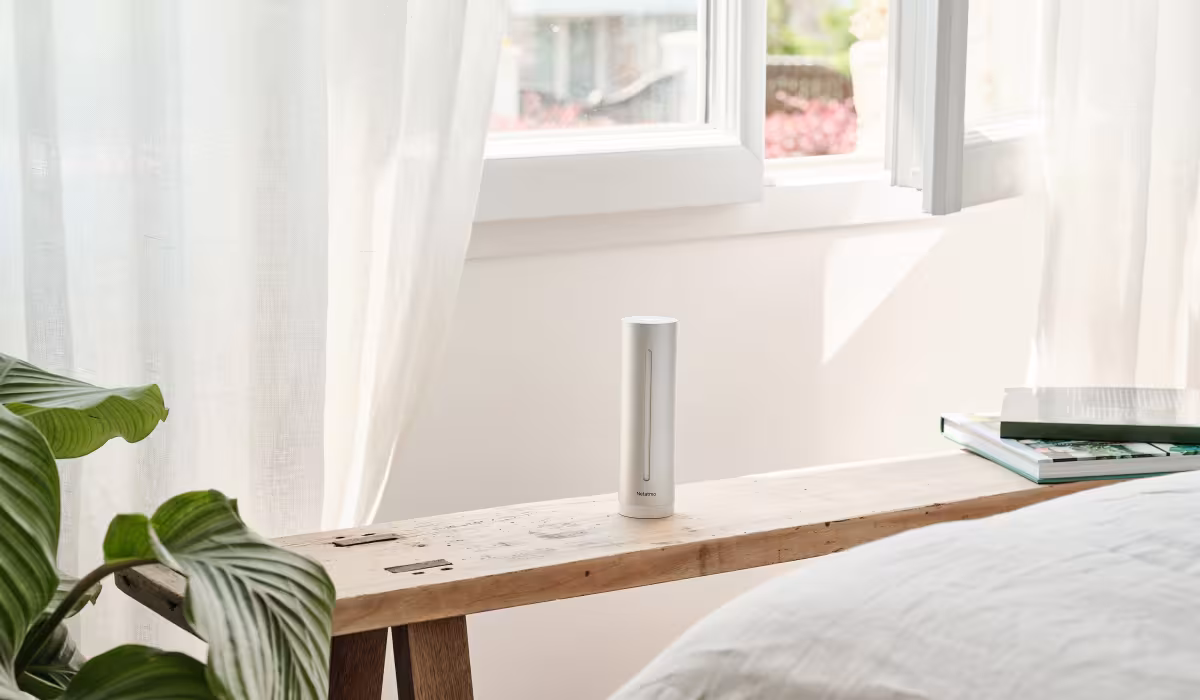 Stazione meteo intelligente Netatmo su panca in legno accanto a una finestra luminosa con tende bianche e pianta verde