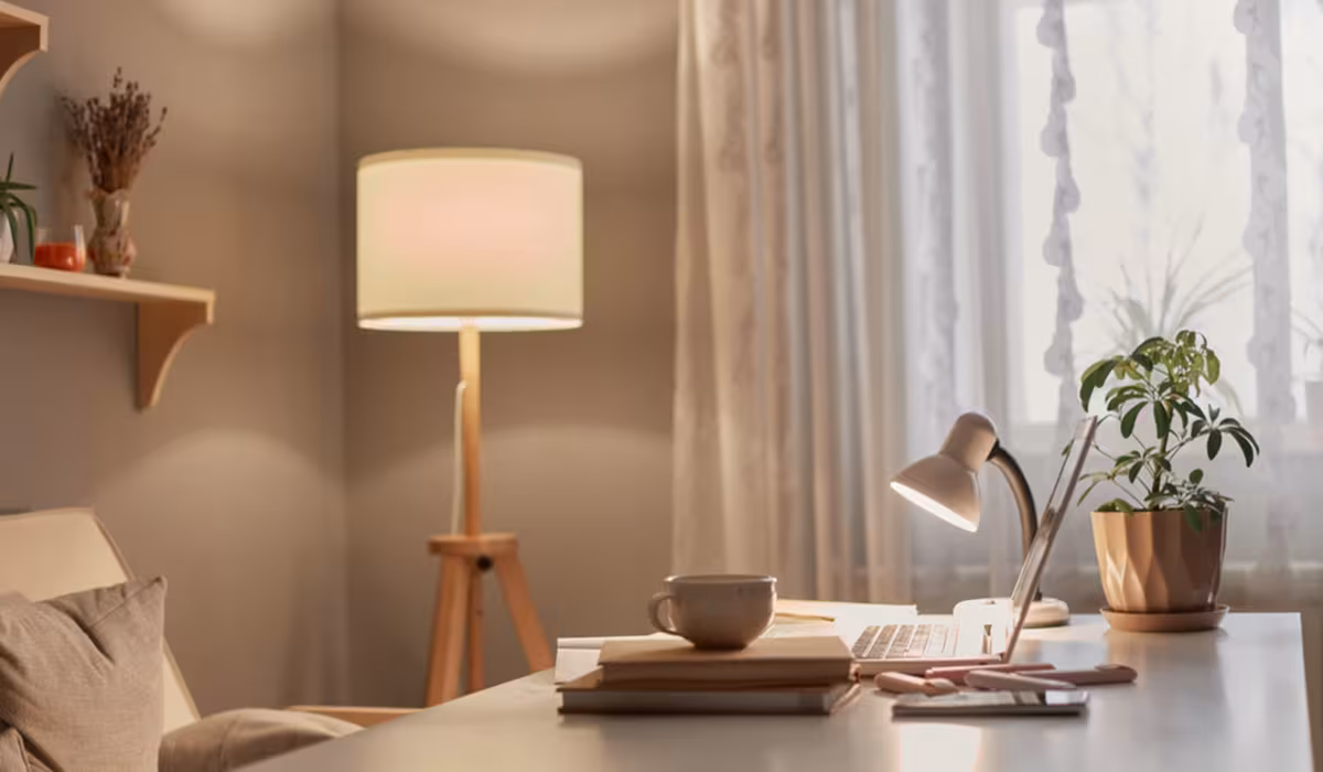 Salotto accogliente con lampada da terra accesa e scrivania illuminata da una lampada da tavolo, con laptop, tazza e pianta verde