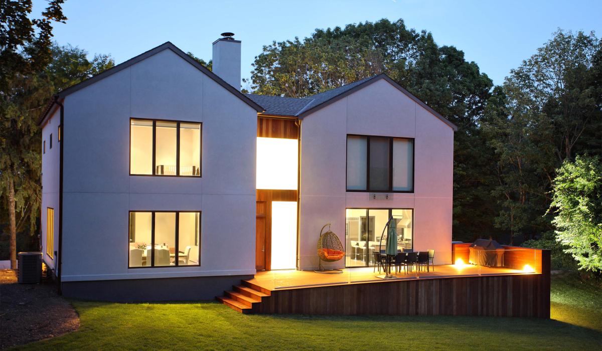 Casa moderna a due piani con ampie vetrate illuminate, terrazza in legno con arredi da esterno al tramonto