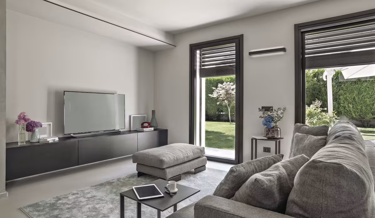 Soggiorno con divano grigio, tavolino con tablet e tazza, TV su mobile e infissi con tapparelle automatizzate affacciate sul giardino