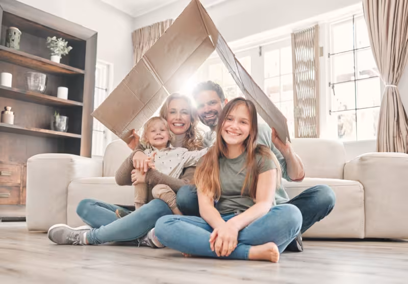 Famiglia sorridente seduta sul pavimento del soggiorno con un cartone sopra la testa, come simbolo di protezione e gioco