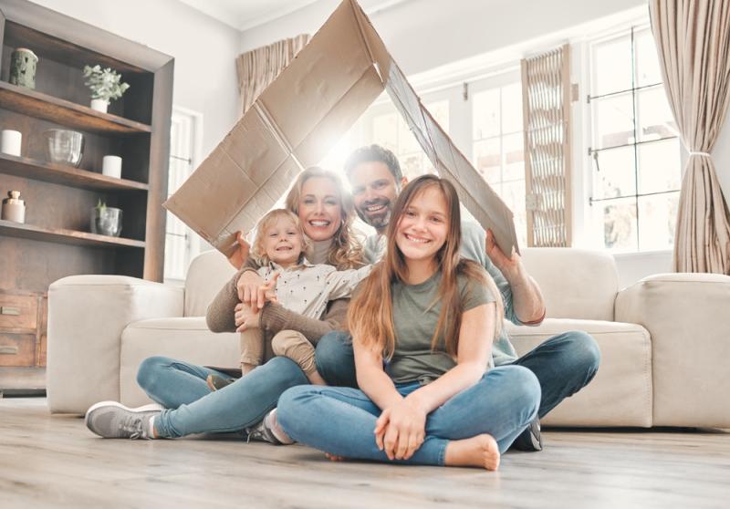 Famiglia sorridente seduta sul pavimento del soggiorno con un cartone sopra la testa, come simbolo di protezione e gioco