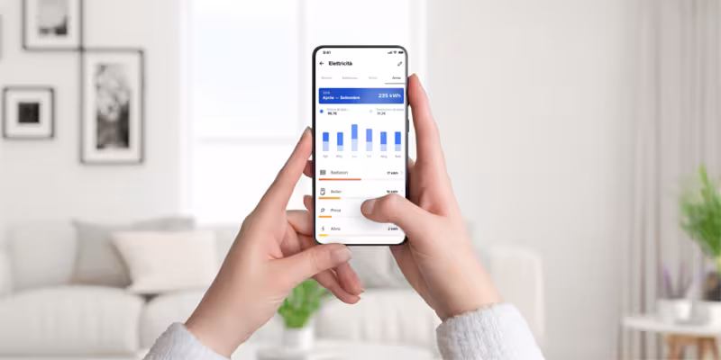 Mani che tengono uno smartphone con schermata dell’app Home + Control di Bticino per il monitoraggio dei consumi elettrici, in un salotto moderno