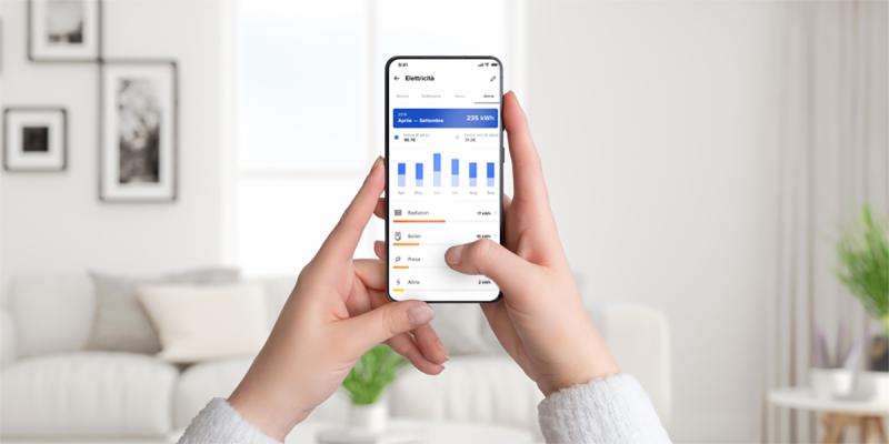 Mani che tengono uno smartphone con schermata dell’app Home + Control di Bticino per il monitoraggio dei consumi elettrici, in un salotto moderno