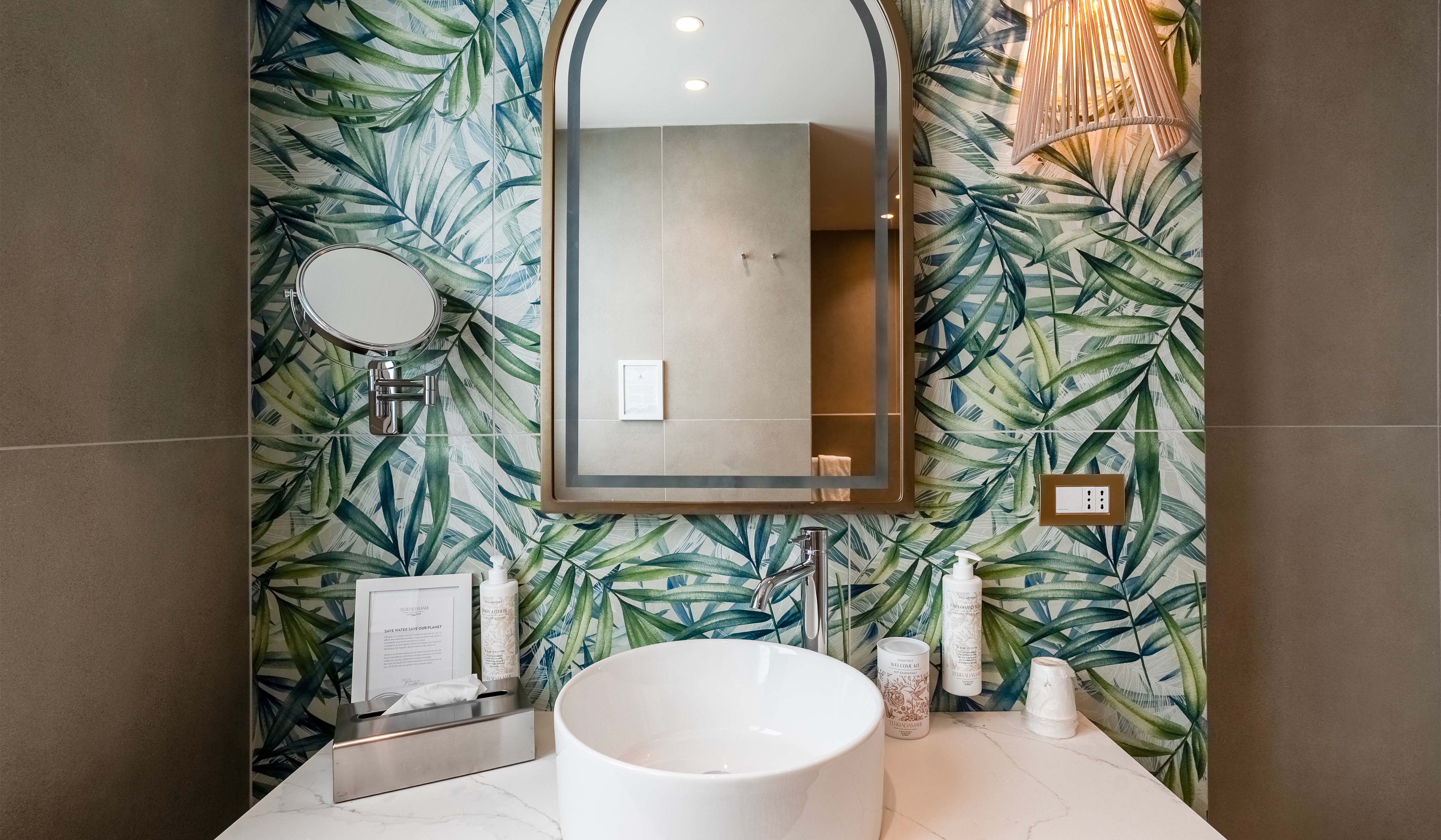 Bagno Terradamari Resort & Spa Bagno del Terradamari Resort & Spa con lavabo moderno, specchio ad arco, parete decorata con motivi tropicali e placche Livinglight Bticino