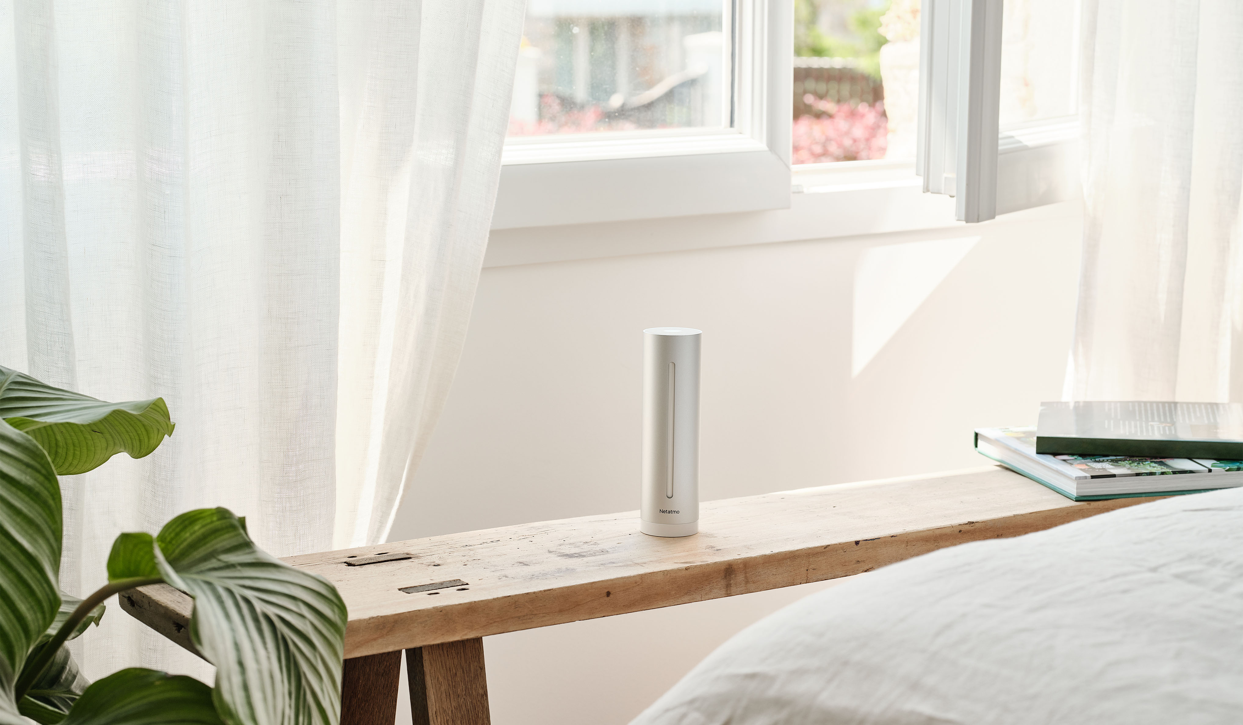 Stazione meteo intelligente Netatmo su panca in legno accanto a una finestra luminosa con tende bianche e pianta verde