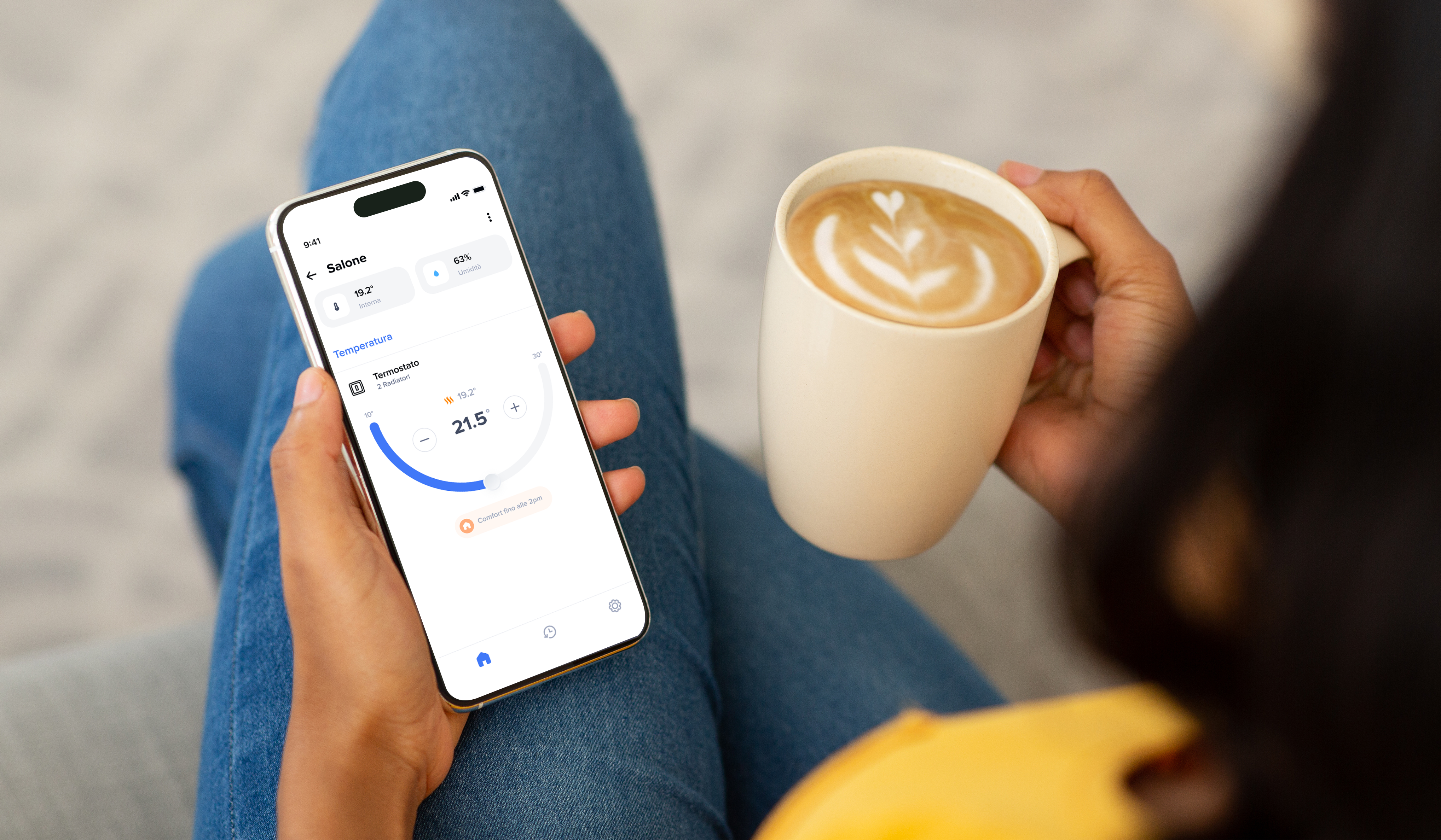 Donna seduta con smartphone con schermata app Home + Control per la gestione del termostato mentre tiene una tazza di caffè in mano