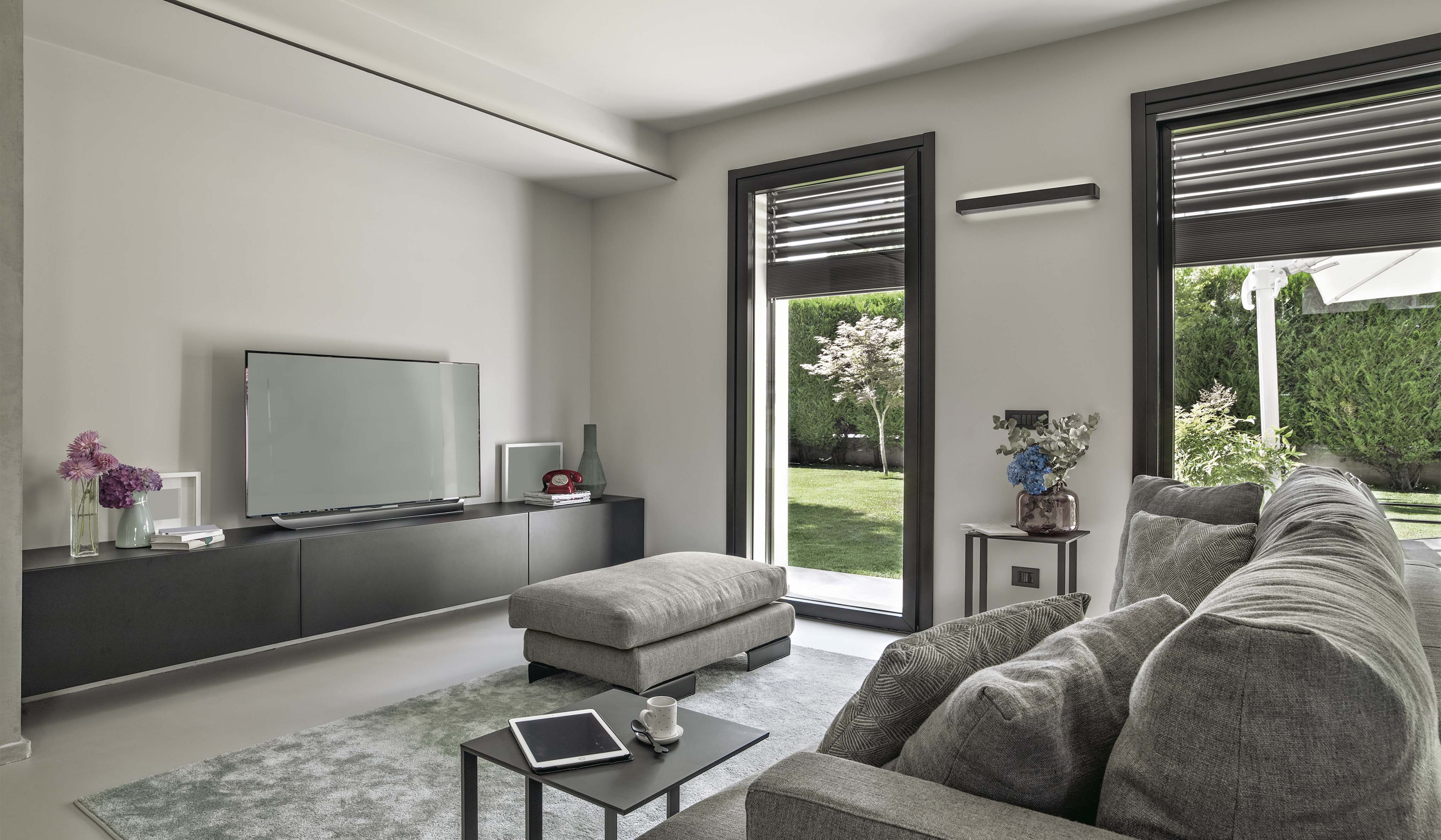 Soggiorno con divano grigio, tavolino con tablet e tazza, TV su mobile e infissi con tapparelle automatizzate affacciate sul giardino